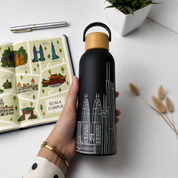A hand holds a matte black water bottle with a light wood cap and white Kuala Lumpur cityscape line art. An illustrated open book, a pen, and dried stems are arranged on a white surface.