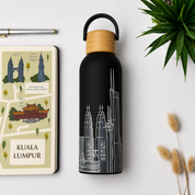 A matte black water bottle with a light wood cap and black loop handle, printed with a white outline of the Kuala Lumpur skyline. It is styled in a flat-lay arrangement next to a travel notebook, a pen, and a green plant on a white background.