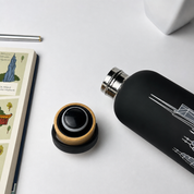 A matte black insulated bottle with a silver metal cap, shown beside its open lid on a light gray surface. The lid has a natural wood ring and a black center.