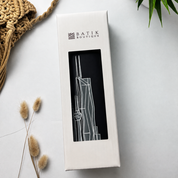 A tall, narrow white product box with a clear window showing a black insert featuring a minimalist white line drawing of a skyscraper. The box is branded 'BATIK BOUTIQUE' and is photographed on a light gray surface with decorative dried grass stems, a woven textile, and a green plant leaf.