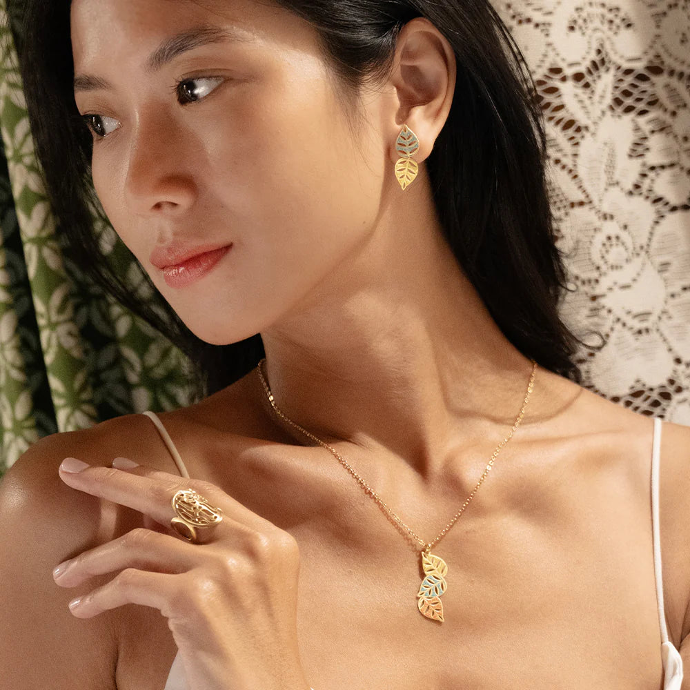 Woman wearing gold jewelry including earrings, necklace, and ring with a lace curtain background