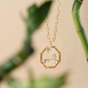 Gold necklace with a hexagonal pendant featuring a bird and leaves against a blurred green background