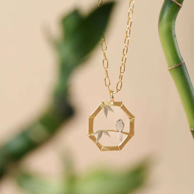 Gold necklace with a hexagonal pendant featuring a bird and leaves against a blurred green background