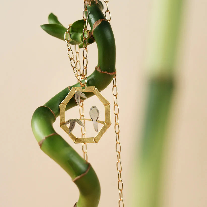 Gold necklace with a hexagonal pendant featuring a bird design, hanging on a bamboo branch.