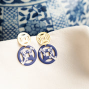 A pair of dangling earrings displayed on a cream fabric surface, with a blue-and-white patterned ceramic bowl in the background. Each earring has a gold-tone circular top with a cutout symbol and a navy blue circular drop featuring a white floral branch motif.