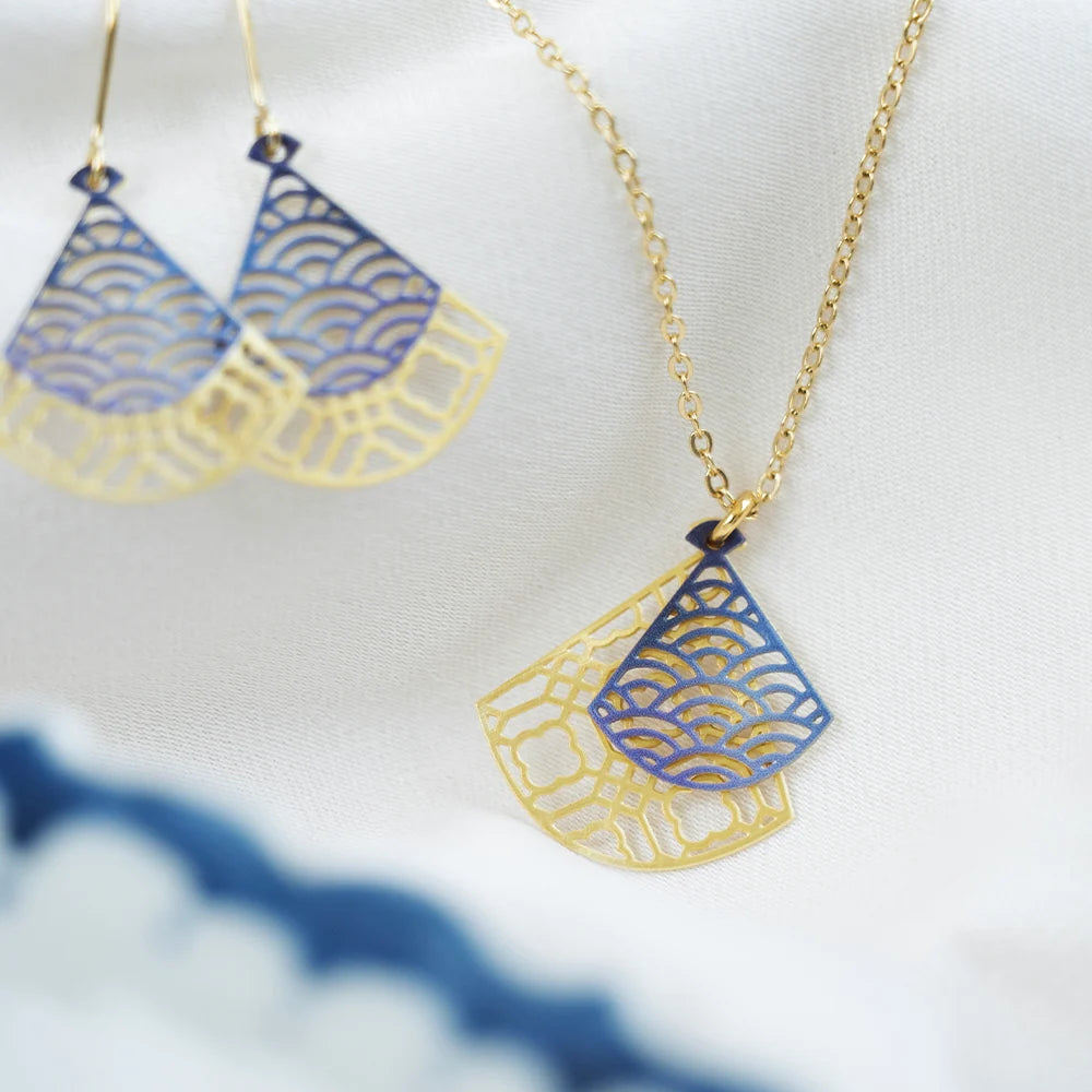 Gold-tone necklace and matching drop earrings with fan-shaped cutout pendants in blue and gold, displayed on a soft white fabric background.