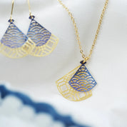 Gold-tone necklace and matching drop earrings with fan-shaped cutout pendants in blue and gold, displayed on a soft white fabric background.
