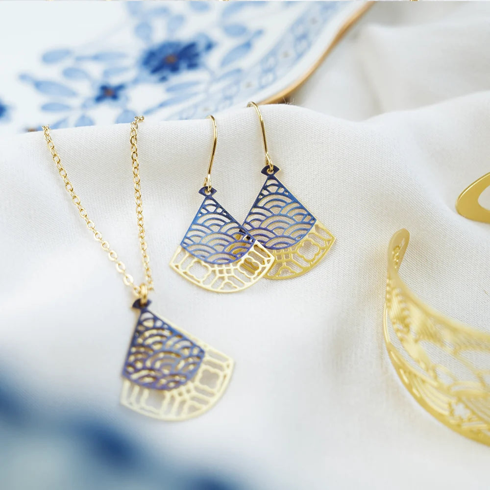 Gold-tone jewelry set featuring a necklace and matching earrings with fan-shaped cutout pendants decorated with blue wave and geometric patterns, displayed on a soft white fabric.