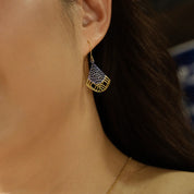 A close-up of a woman's ear wearing a gold-toned dangle earring with a blue and white wave pattern, paired with a delicate gold chain necklace.