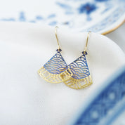 Gold and blue fan-shaped dangle earrings with a wave pattern, resting on a white textured fabric next to a blue and white ceramic plate.