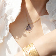 A gold necklace with a geometric pendant featuring a dark purple inlay, worn by a model in a white lace top. A matching gold cuff bracelet is also visible.