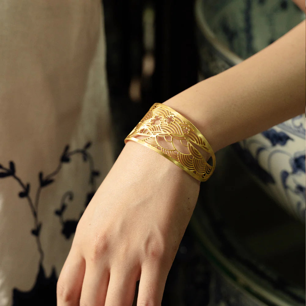 A close-up of a person's wrist wearing a wide, gold-toned cuff bracelet with an intricate openwork floral pattern.