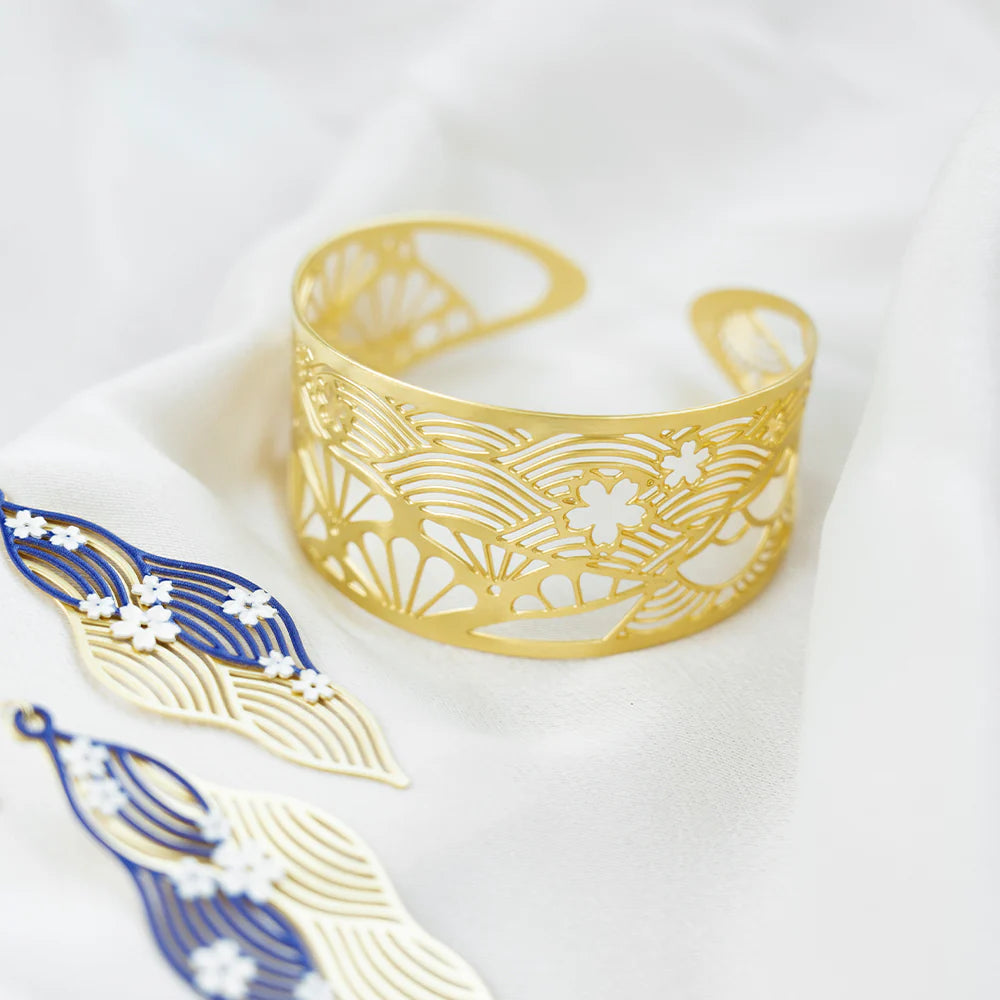 A gold-toned open cuff bracelet with an intricate wave and cherry blossom cutout pattern, displayed on draped white fabric alongside matching blue and gold leaf-shaped earrings.