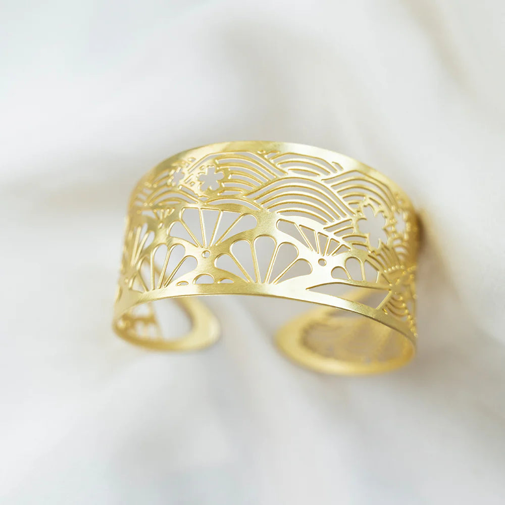 A gold-colored open cuff bracelet with an intricate laser-cut fan and wave pattern, resting on a soft white fabric background.