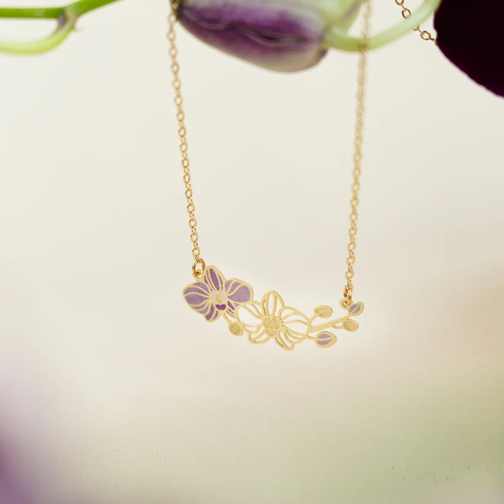 A delicate gold chain necklace with a floral pendant featuring purple and gold orchid details, shown against a soft, blurred background with a purple flower stem at the top.