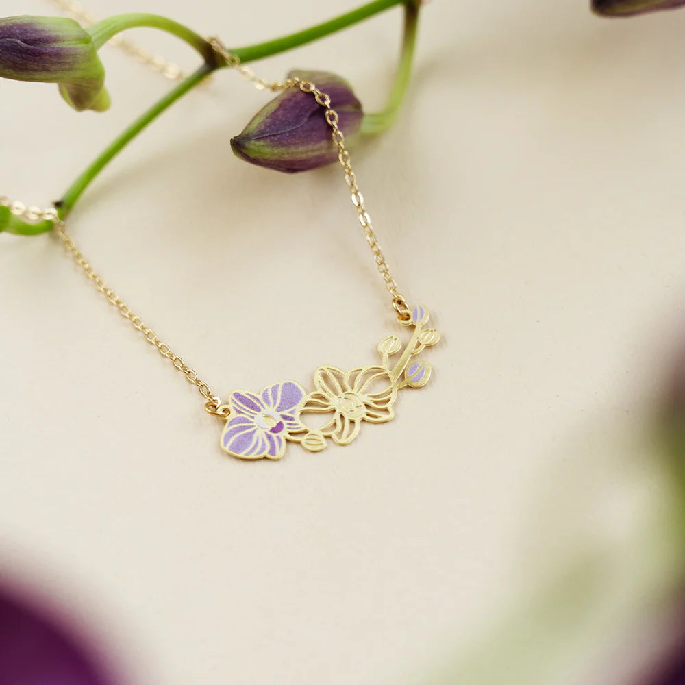 A delicate gold chain necklace featuring three small floral charms, including one lavender-purple orchid flower, displayed on a soft cream background with blurred purple flowers.
