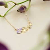A delicate gold chain necklace featuring three small floral charms, including one lavender-purple orchid flower, displayed on a soft cream background with blurred purple flowers.