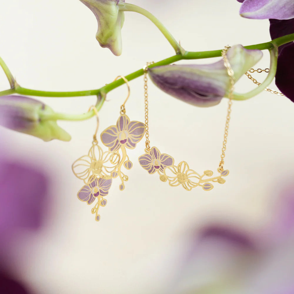 Gold-tone orchid flower dangle earrings and a matching chain necklace displayed on a green orchid stem with soft purple blossoms in the background.