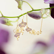 Gold-tone orchid flower dangle earrings and a matching chain necklace displayed on a green orchid stem with soft purple blossoms in the background.