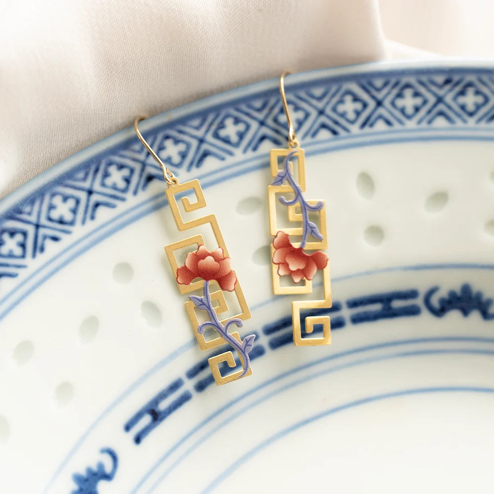 A pair of gold-tone drop earrings with geometric filigree and pink enamel flowers with purple stems, displayed on a blue and white ceramic plate.