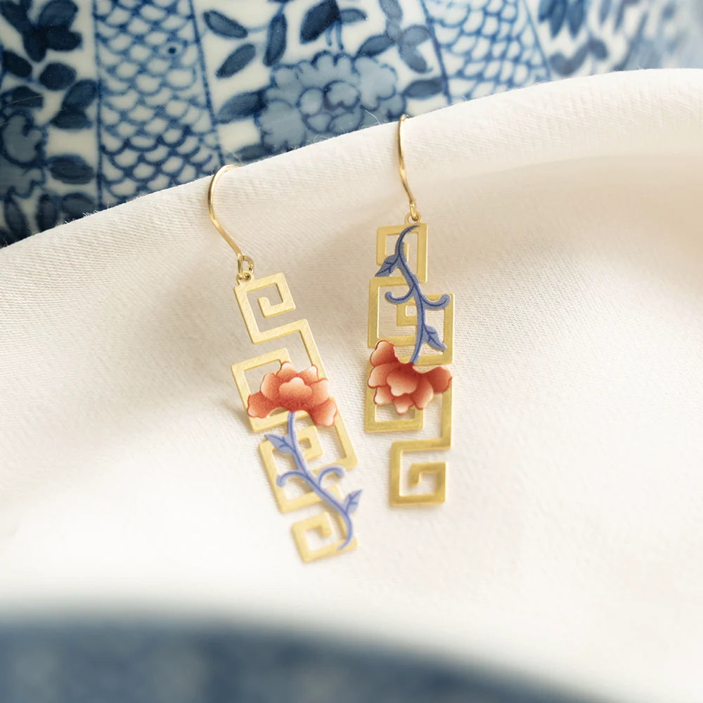 A pair of gold-tone drop earrings with geometric cutout frames, decorated with pink floral motifs and blue vine details, resting on a white textured surface.