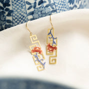 A pair of gold-tone drop earrings with geometric cutout frames, decorated with pink floral motifs and blue vine details, resting on a white textured surface.