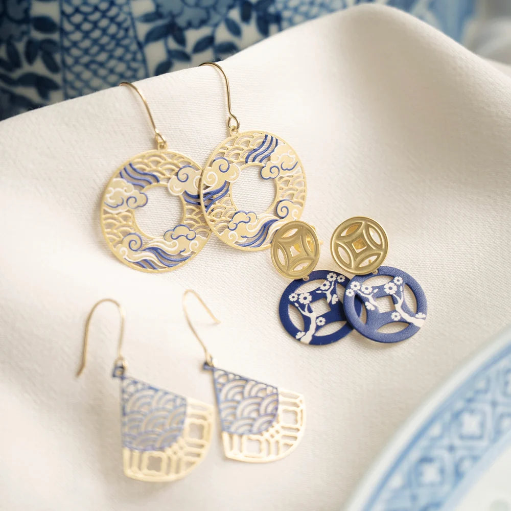 A styled flat-lay of gold and blue Japanese-inspired earrings arranged on a white textured fabric with a blue patterned ceramic plate edge in the background.