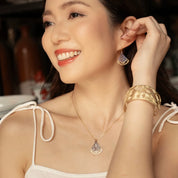 A woman wearing a delicate gold necklace with a geometric pendant, a matching gold cuff bracelet, and a gold hoop earring with a purple crystal accent, posing in a warm indoor setting.