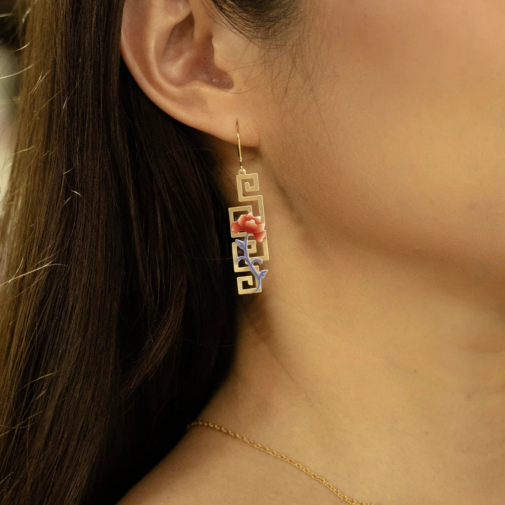 A close-up of a woman's ear and neck wearing a gold Greek key dangle earring with a coral-pink and blue floral accent, paired with a delicate gold chain necklace.