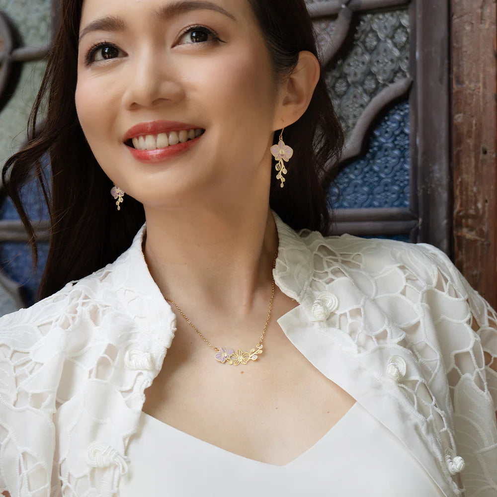 A woman wearing delicate gold jewelry, including a floral necklace and matching earrings, paired with a white lace-trimmed top. She is smiling and looking off-camera against a rustic wooden and blue-gray background.