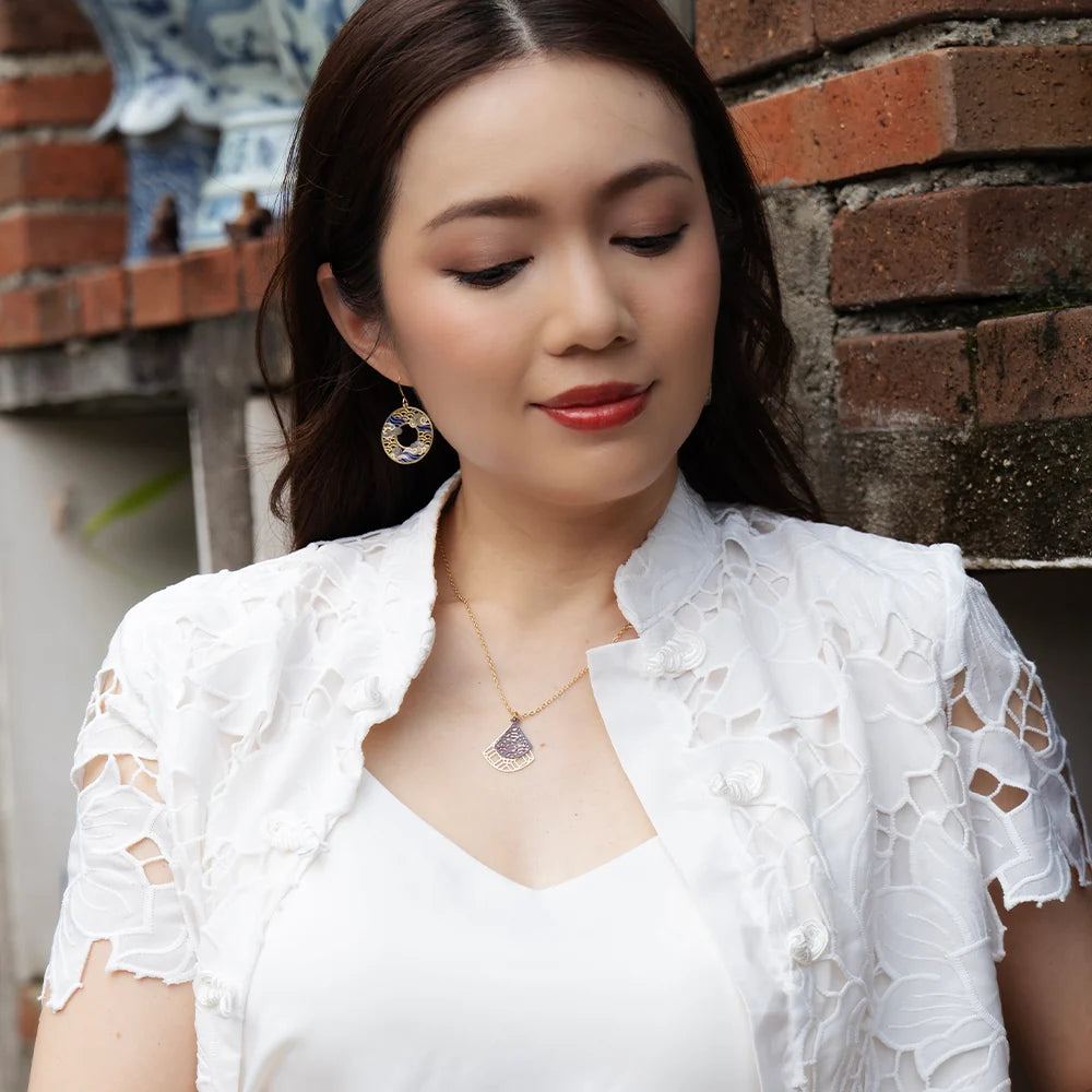 A woman wearing a white lace short-sleeve cardigan over a white top, accessorized with a gold pendant necklace and matching earrings, standing against a brick wall.