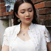 A woman wearing a white lace short-sleeve cardigan over a white top, accessorized with a gold pendant necklace and matching earrings, standing against a brick wall.