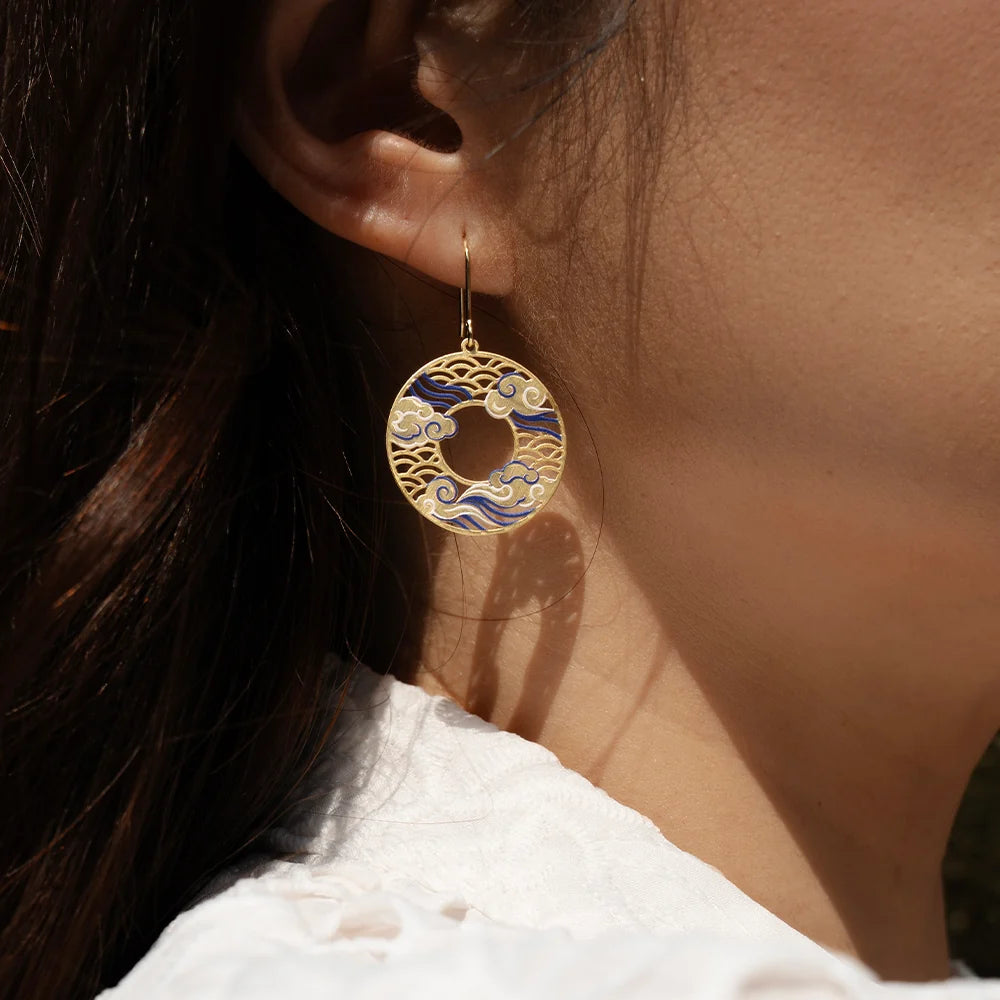 A close-up of a person's ear wearing a dangling circular earring with gold-tone metal and blue-and-white wave patterns, against warm sunlight.