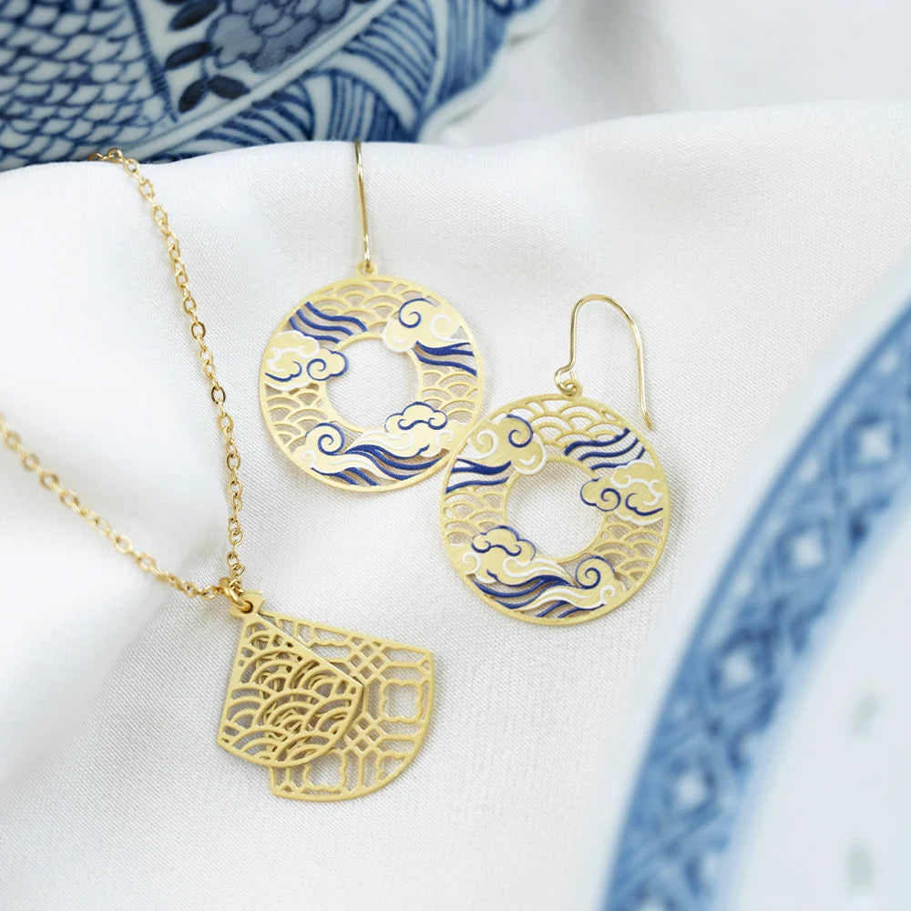 Gold jewelry set featuring a necklace and matching earrings with blue and white cloud-patterned enamel details, displayed on a white fabric surface with a blue patterned ceramic plate in the background.