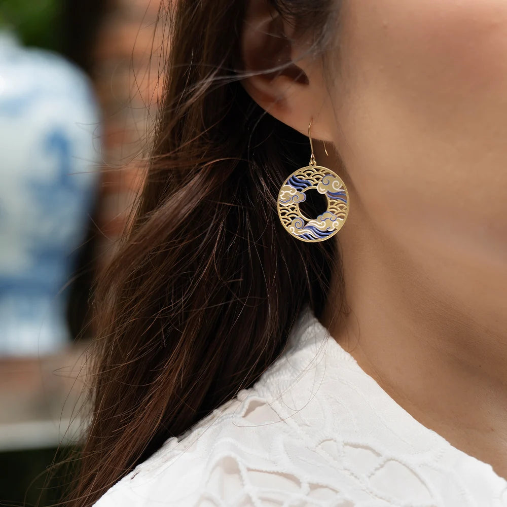 A close-up of a person wearing a dangling circular earring with gold filigree and blue wave-like enamel details, paired with a white textured top.