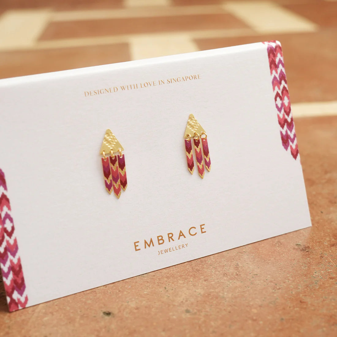 Gold and red earrings on a 'Embrace Jewellery' card with a wooden surface background