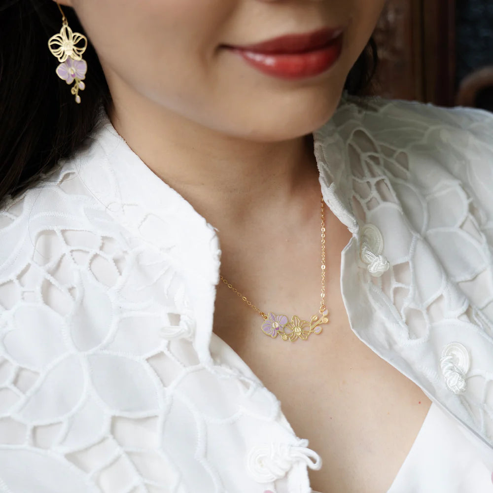 A close-up lifestyle shot of a woman wearing a delicate gold necklace with a butterfly and flower pendant, along with a matching butterfly earring. She is wearing a white lace blouse.