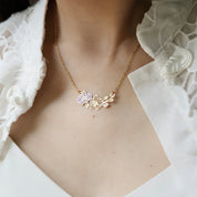 A gold chain necklace with a delicate pendant featuring a lavender flower and gold wire floral details, worn on a model's neck over a white lace blouse.